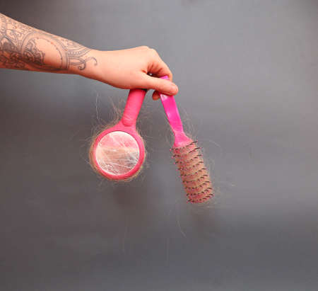 Hair loss in comb. female hand with a tattoo holds a pink comb with white hair on a gray background. Hair health conceptの写真素材