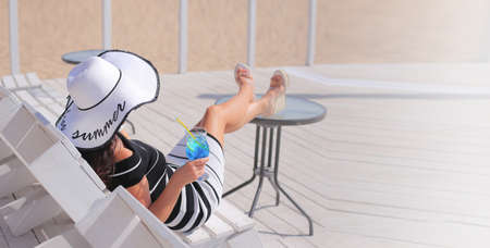 A beautiful woman sits at a table in a beach bar with a straw hat on her head drinking a cocktail Copy space.の写真素材