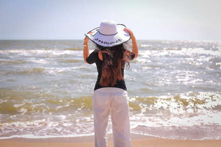 beautiful woman in sunhat white pants, black blouse, standing with her arms raised to her head on summer day.の写真素材