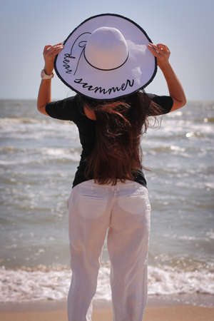 beautiful woman in sunhat white pants, black blouse, standing with her arms raised to her head on summer day.の写真素材