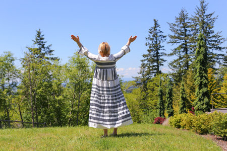 A woman in a white dress with white hair stands facing the forest with her hands up, praise and delightの写真素材