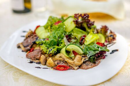 warm salad with meat, cucumber and vegetables on a white plate on the tableの写真素材