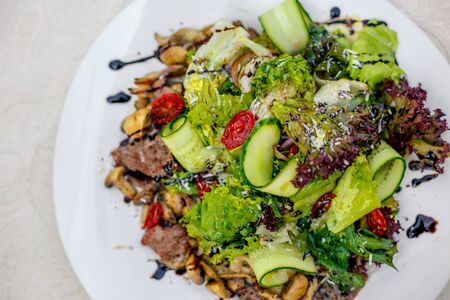 warm salad with meat, cucumber and vegetables on a white plate on the tableの写真素材