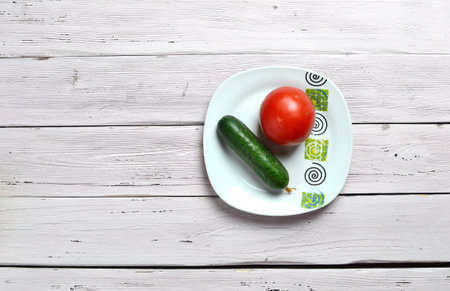 still life with one tomato and one cucumber on plate with ornament isolated on an old wooden light rustic tableの写真素材