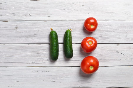 close up of three red tomatoes and two green cucumbers on an old wooden light rustic tableの写真素材