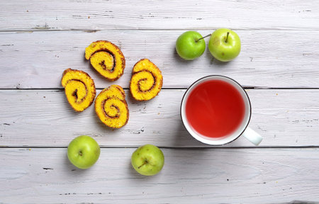top view of delicious biscuit rolls with apricot jam, green apples and cup of red drink on a white rustic wooden tableの写真素材