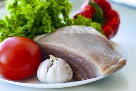 A piece of raw pork on a white plate with lettuce, sweet pepper, tomato and garlic. Healthy eating and cooking at home conceptの写真素材