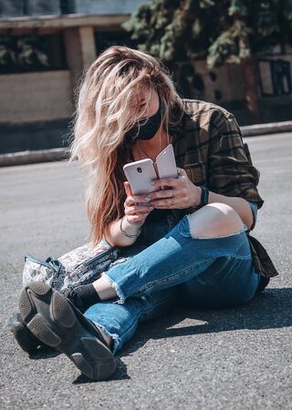 Young blonde girl in black protective medical mask is looking into phone screen.の写真素材