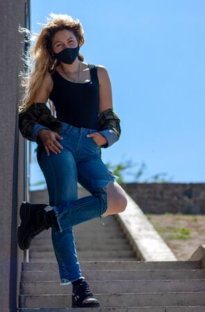 Girl in black protective medical mask  stands near black wall.She is dressed in black T-shirt, a plaid shirt and jeans.の写真素材