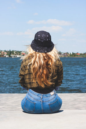 Girl sits near the river in a sunny day.She is dressed in    a plaid shirt and blue hatの写真素材
