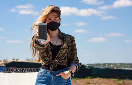 Girl in black protective medical mask is holding a phone with black screen.She is dressed in black T-shirt, a plaid shirt and jeans. Social media and news concept.  Mock upの写真素材