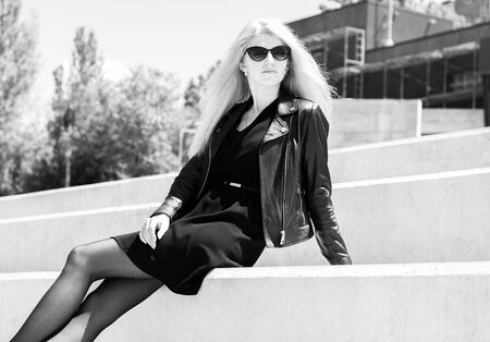 A girl with black "cat-eye" glasses wears a black dress and leather jacket looks far away. She sits on the steps outside in the city in a sunny day. Black and white photoの写真素材