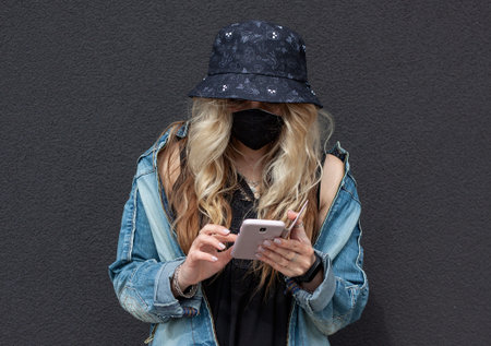 Young blonde  girl in a black protective mask, blue hat, denim jacket and black shirt is making a...