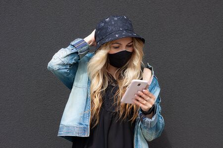 Young blonde  girl in a black protective mask, blue hat, denim jacket and black shirt is checking social apps. Concept of Corona-virus isolation and communication. She stands near black wallの写真素材