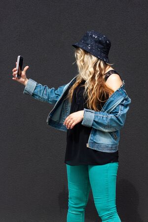 Young blonde  girl in a black protective mask, blue hat, denim jacket and black shirt is making a selfie. Concept of Corona-virus isolation and communication. She stands near black wallの写真素材
