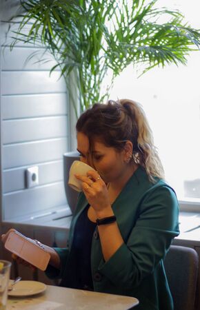 Young blonde girl in a cafe in a green jacket drinks coffee and looks at the phoneの写真素材