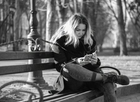 Black and white photo of a girl in a coat in a park sits in an interesting pose on a bench with a phone in her hands. Social Media Conceptの写真素材