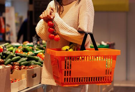Girl holds a branch of tomatoes in her right hand and an orange basket  full of fruits in her left hand in a supermarket.の写真素材