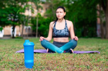 The bottle of water is the main object on the photo. There is a blurred girl is sitting on the karemat and doing yoga の写真素材
