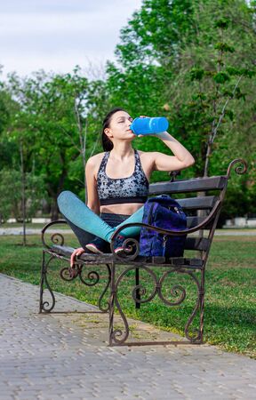 Beautiful  brunette girl with a backpack   is  sitting on the bench  in the park and drinks water aster training.  Concept of sport の写真素材