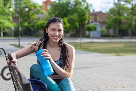 Beautiful smiling brunette girl  is sitting on the bench  in the park and drinks water aster training.  Concept of sport の写真素材
