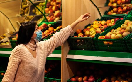 Beautiful brunette girl is  taking a yellow apple in the supermarket. She is wearing light sweater, white jeans and blue medical maskの写真素材