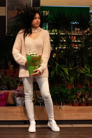 Beautiful brunette  girl is holding a   pot with white flowers in the supermarket. She is wearing light sweater and white jeansの写真素材