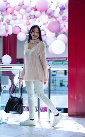Beautiful  girl is walking in the shopping center. She is wearing light sweater and white jeans and holding a black bagの写真素材