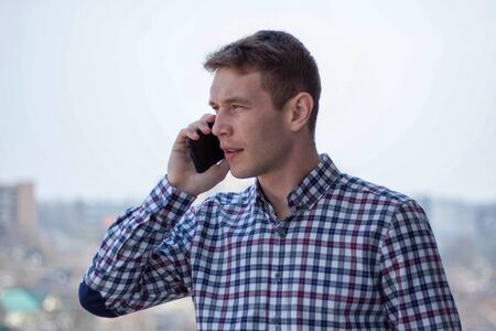 Caucasian  men model is making a business call. There is a city on the back ground.  Dressed into checkered shirt. Stands on the 9th floor.の写真素材