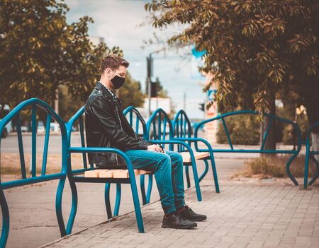 Caucasian sad men sits on the bench and looks straight away. He is depressed. Dressed into jeans, T-shirt, leather jacket and black protective mask.  (Picture has a preset dark colors.)の写真素材