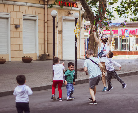 Nikolaev, Ukraine- June 6, 2020: a tall bearded man makes a big soap bubble on the street. 5 happy children run around him and have fun on the main city street Sobornaのeditorial素材