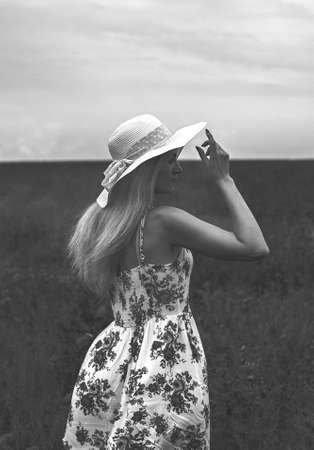 Beautiful girl in a white summer hat is dreaming in a poppy field. She is dressed in a white dress with red flowers (black and white photo)の写真素材