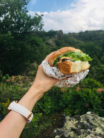 A girl holds a bite burger in her hand against the background of a beautiful landscape of nature.の写真素材