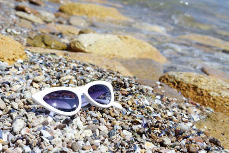White female glasses called "cat's eye" on the stones, pebbles and sand by the sea water. Leisure and vacation concept. Fashion style retro of 80's last centuryの写真素材