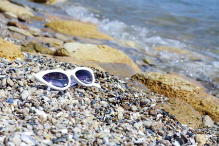 White female glasses called "cat's eye" on the stones, pebbles and sand by the sea water. Leisure and vacation concept. Fashion style retro of 80's last centuryの写真素材