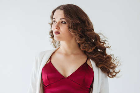 A beautiful curly pregnant girl in a dark pink evening dress and long white jacket is on a white background. (selective focus on one eye)の写真素材