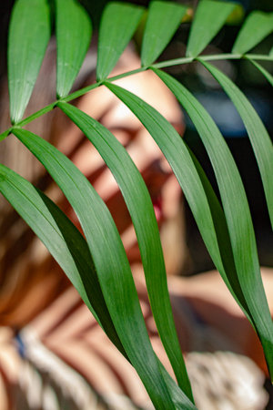 A very beautiful woman stands outside under a palm leaf. She is blurred but the leaf is clear into...