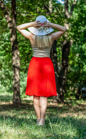 A very beautiful woman stands in the park (picture from the back side). She is dressed into white hat and light top, red skirt. She is in the park, the weather is warm sunny summerの写真素材