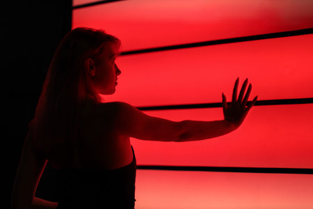 Pretty sexy young girl dancer in fashionable youth clothes stands near neon red wall and touches it...