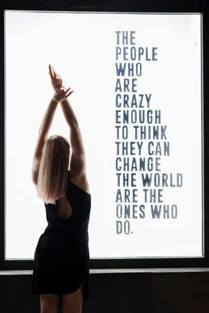 girl in dress stands with raised up and crossed hands near white neon wall at night. It is written on the wall: the people who are crazy enough to think they can change the world are the ones who do.の写真素材