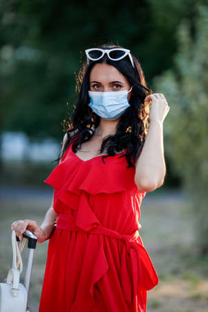 Young brunette woman with cat-eyes shaped sunglasses in a madical mask stands outside with a suitcase. It is hot weather outside. Conceptual of covid and protectionの写真素材