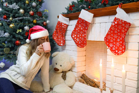 Young lady in white sweater drinking from cup near fireplace. New Year tree and home decoration. Candles and teddy bear, gifts are around. Concept of Christmas holidays. Calm and peaceful atmosphereの写真素材