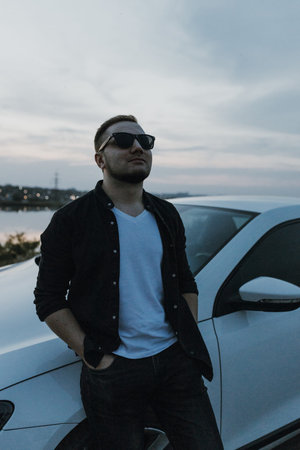 Handsome man in black T-shirt and white shirt with jeans and sneakers poses near modern white sport car and looks at the sunset outdoors. Fresh weekend out of the city near riverside. dark tone photoの写真素材