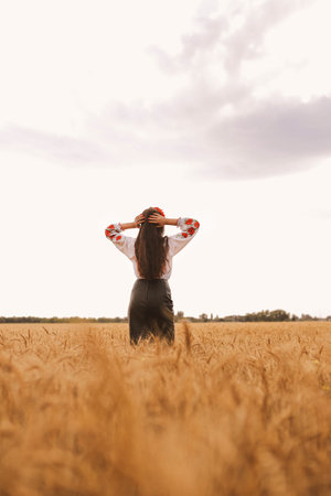 Young pretty girl stands in the field wearing Ukrainian national embroidered shirt with red wreath....