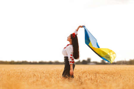 Young pretty girl holds flag and waiting for victory in the field wearing Ukrainian national embroidered shirt with red wreath. Concept of Ukraine Independence, freedom and stop the war. peace in wordの写真素材