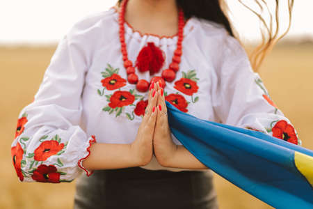 Young girl prays in the wheat field wearing Ukrainian national embroidered shirt with flower red wreath and holds flag at sunset yellow light. Concept Independence, freedom of Ukraine and stop the warの写真素材