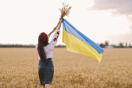 Young girl waiting for victory in the field wearing Ukrainian national embroidered shirt with red wreath and holds up flag and wheat bouquet . Independence Concept, freedom of Ukraine and stop the warの写真素材