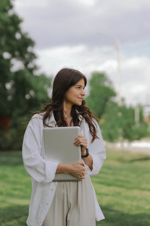 Beautiful Caucasian girl walking with laptop computer on green grass in summer park. Happy female...