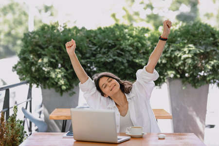 Portrait happy young office manager working remotely online sitting with laptop, tablet and coffee outside on terrace raised hands up after getting big contract. remote freelance successful jobの写真素材