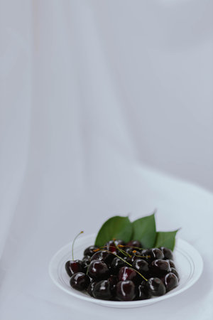 Fresh ripe dark black huge sweet cherries with leaves and water drops on white plate, light cloth...